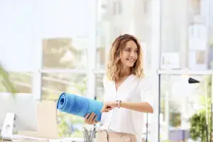 Smiling wellness consultant holding a yoga mat in a bright, modern workspace, symbolising holistic health and leadership wellbeing.