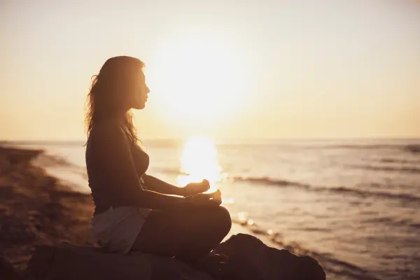 Professional woman meditating near ocean at sunrise, reflecting calm focus and balance in leadership.